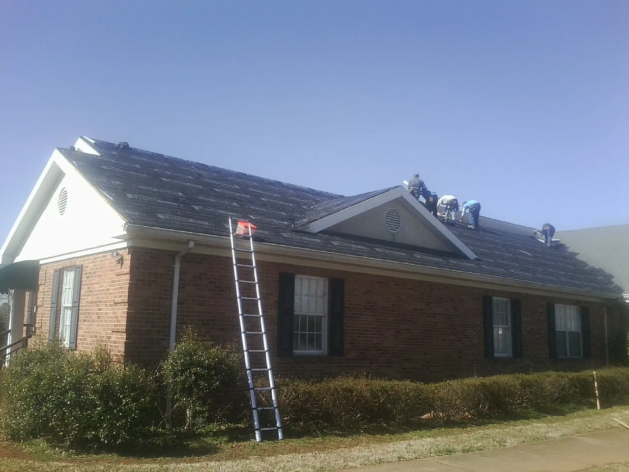 Crew tearing shingles off a brick ranch roof with a ladder set — Emergency Roof Repair work by our crew in Galeton