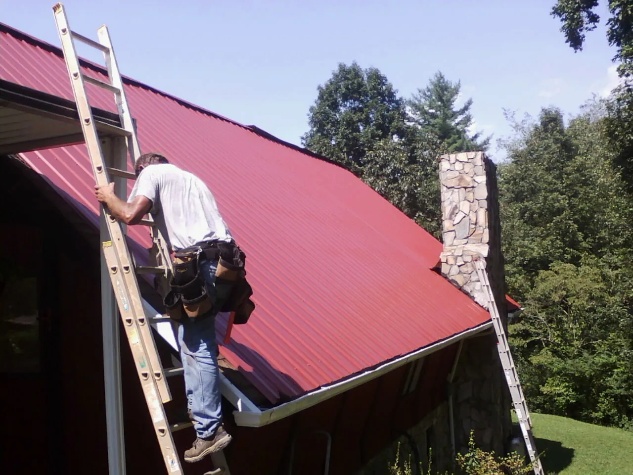 Roofer on a ladder beside a red metal roof and stone chimney — Soffit & Fascia Repair by Roofing Brothers in Galeton